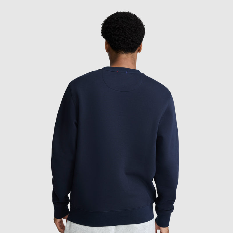 A person with short curly hair is wearing a navy blue Champion Powerblend Crewneck Sweatshirt with a tonal script logo and light gray pants, standing with their back to the camera against a plain light gray background.