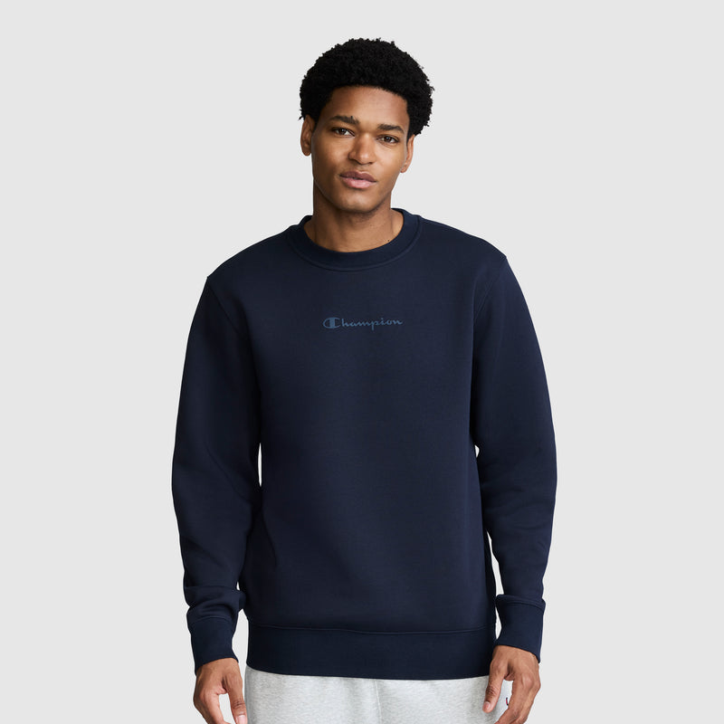 A man wearing a Champion Powerblend Crewneck Sweatshirt with a tonal script logo in navy blue and light gray pants stands against a plain light gray background, looking at the camera with a neutral expression.