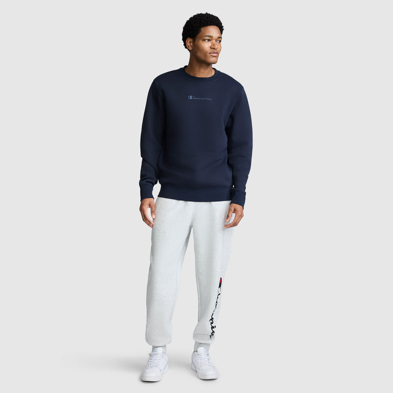 A man stands against a plain light gray background wearing a Champion Powerblend Crewneck Sweatshirt with a tonal script logo in navy blue, paired with light gray sweatpants featuring a logo on the left leg, and white sneakers.
