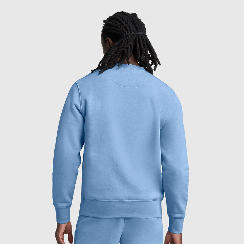 A person with long, dark, braided hair is shown from the back, wearing a light blue Champion Powerblend Crewneck Sweatshirt with a tonal script logo and matching sweatpants against a plain, light gray background.