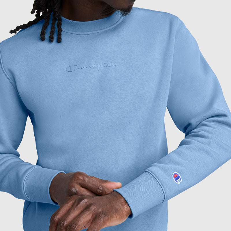 A person wearing a light blue Champion Powerblend Crewneck Sweatshirt, Tonal Script Logo, with subtle embroidered branding on the chest and sleeve, adjusts their sleeve while facing slightly away from the camera.