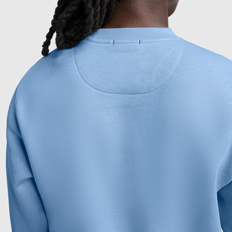A person with dark braided hair is shown from the back wearing the Champion Powerblend Crewneck Sweatshirt, Tonal Script Logo in light blue, featuring visible seams and stitching details on the collar and shoulders.