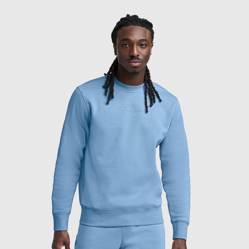 A man with long black hair in twists wears a light blue Champion Powerblend Crewneck Sweatshirt with a tonal script logo and matching pants, standing against a plain white background.