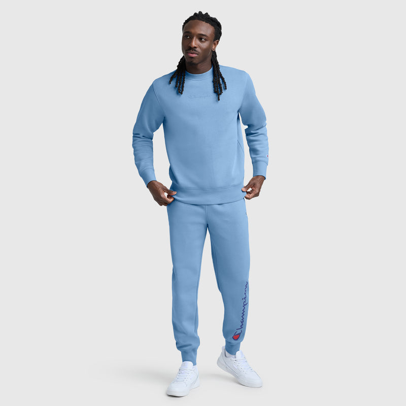 A man with long braids wears a Champion Powerblend Crewneck Sweatshirt featuring a tonal script logo, along with matching light blue sweatpants made from soft fleece. He pairs the outfit with white sneakers and stands against a plain white background.