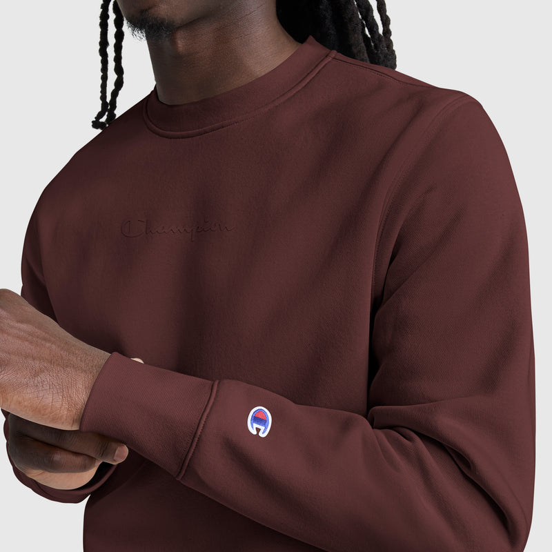 A person wearing the Champion Powerblend Crewneck Sweatshirt in maroon, featuring a tonal script logo embroidered on the chest and a small Champion patch on the sleeve. Crafted from soft Powerblend fleece, only the upper body and arms are visible in this classic men’s sweater style.