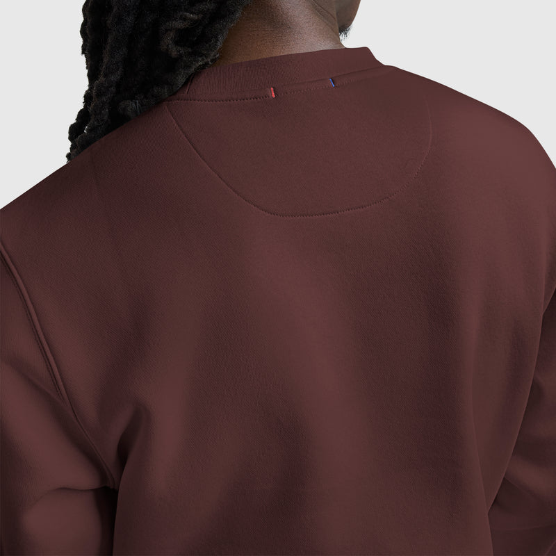 A person with long, dark, braided hair is seen from the back wearing a maroon Champion Powerblend Crewneck Sweatshirt, Tonal Script Logo. The sweatshirt showcases subtle stitching and small red and blue accents near the collar.