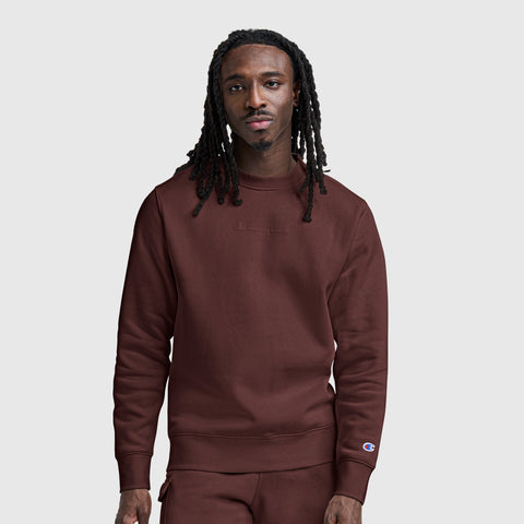 A man with long dreadlocks wears a maroon Champion Powerblend Crewneck Sweatshirt with a tonal script logo and matching pants, standing against a plain light gray background.