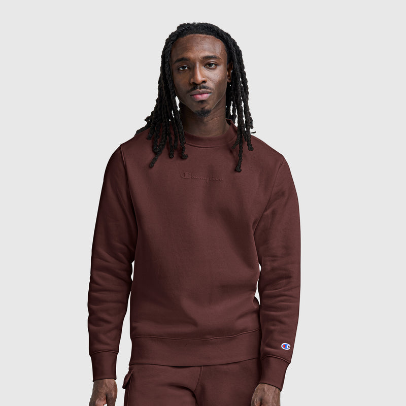 A man with long dreadlocks wears a maroon Champion Powerblend Crewneck Sweatshirt with a tonal script logo and matching pants, standing against a plain light gray background.