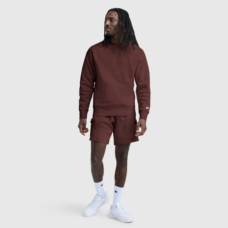 A man stands on a plain gray background wearing a maroon Champion Powerblend Crewneck Sweatshirt with a tonal script logo and matching shorts, paired with white socks and white sneakers. He is looking slightly to his left with one hand relaxed at his side.