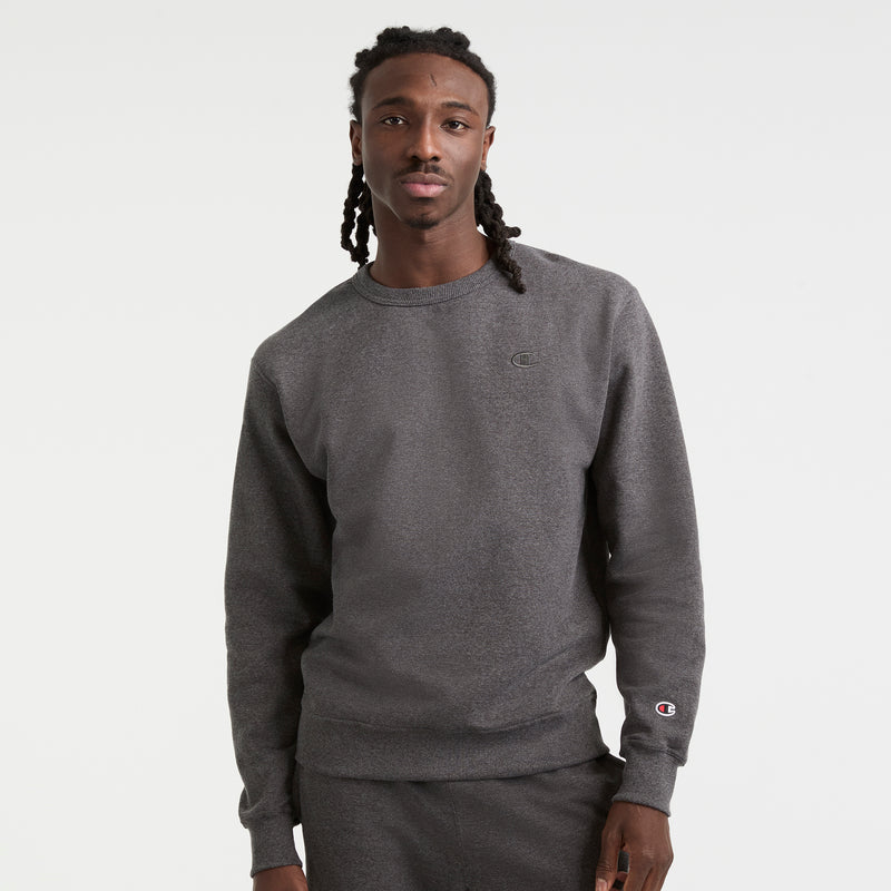 A man with long braided hair wears a dark gray Champion Powerblend Crewneck Sweatshirt, C Logo, paired with matching sweatpants, standing against a plain white background.
