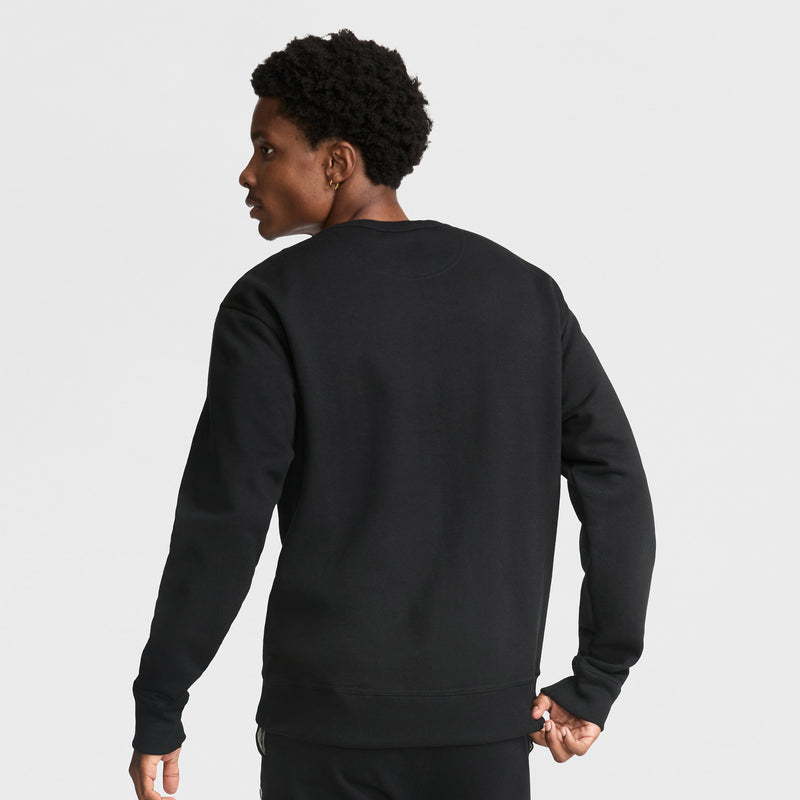 A person with short, curly hair is seen from behind in a plain black Champion Powerblend Crewneck Sweatshirt, C Logo, and black pants, standing against a white background.