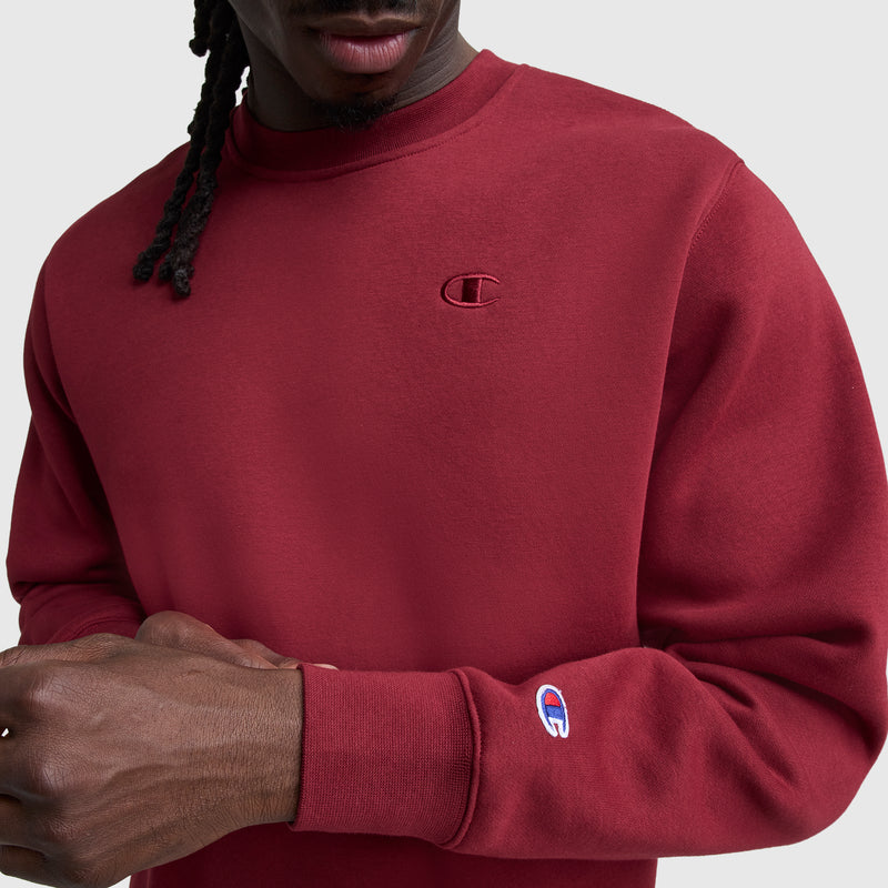 A person wears a maroon Champion Powerblend Crewneck Sweatshirt, C Logo, with chest and sleeve logos, standing against a plain light background. Only their lower face and upper torso are visible.