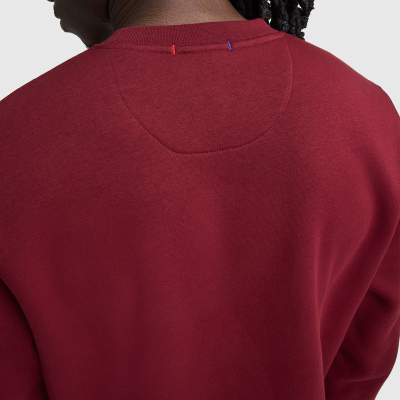 Close-up of the back of a person wearing a plain, dark red Champion Powerblend Crewneck Sweatshirt, C Logo. Only the shoulders, upper back, and part of the neck and head are shown against a light gray background.