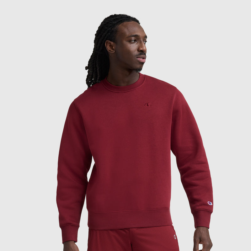 A man with long hair wears the Champion Powerblend Crewneck Sweatshirt, C Logo, in maroon with a small logo on the chest and sleeve. He pairs it with matching shorts and stands against a plain light gray background, looking to the side.