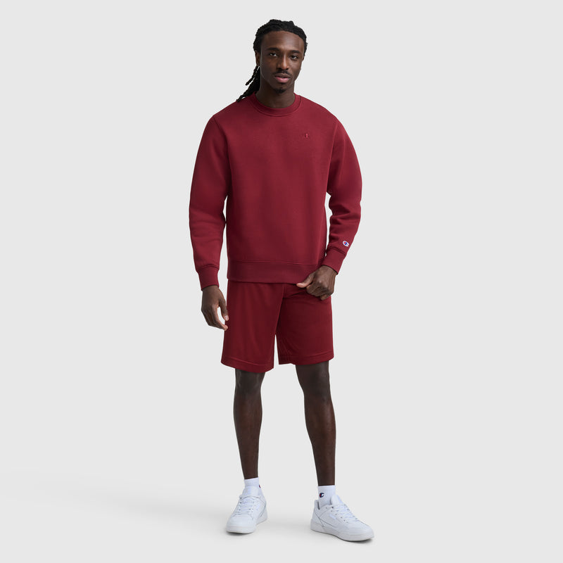 A man with long twists poses confidently against a light gray background, wearing a matching maroon Champion Powerblend Crewneck Sweatshirt with C Logo and shorts set, paired with white sneakers.