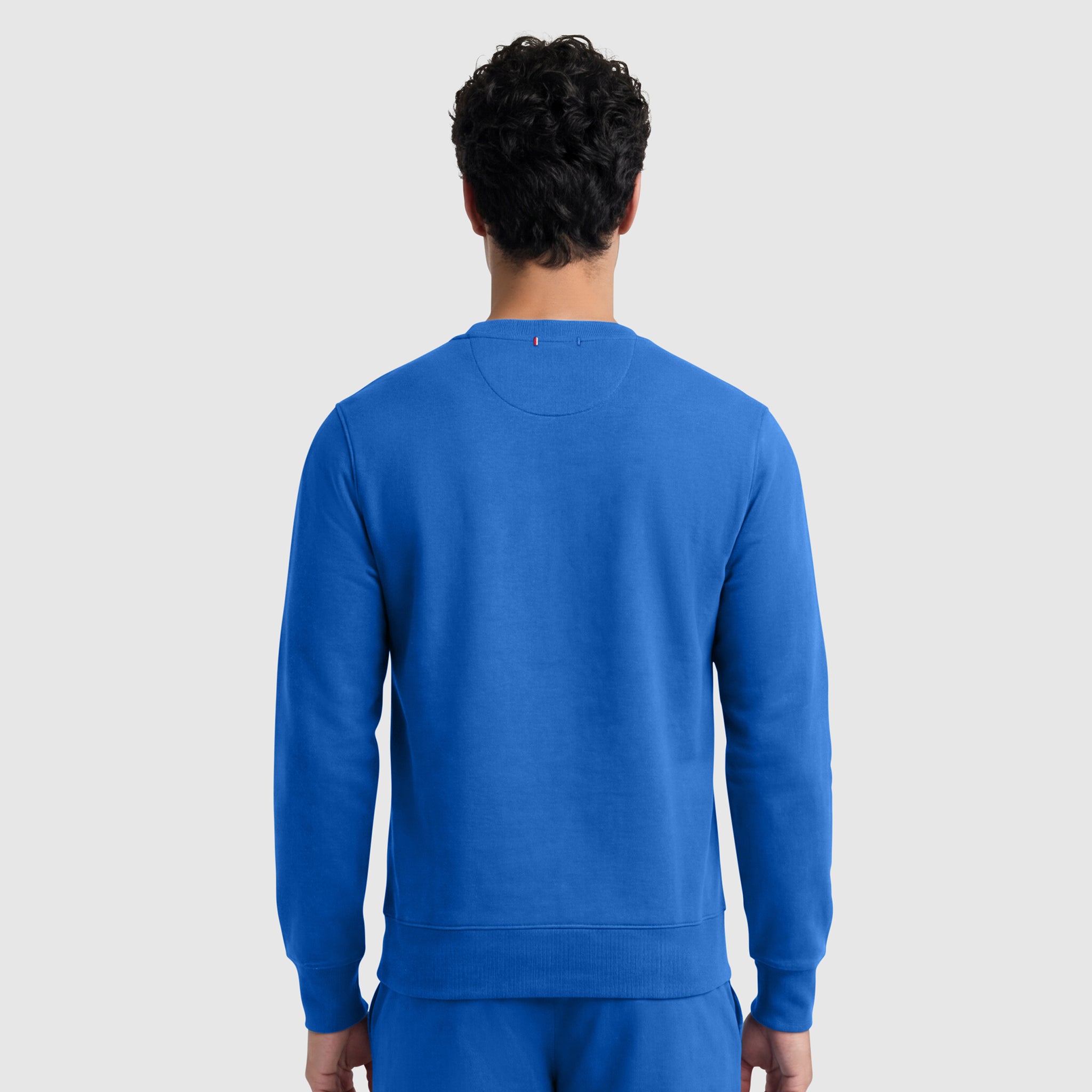 A person with short, curly dark hair is seen from behind wearing the Champion Powerblend Crewneck Sweatshirt, C Logo in bright blue, along with matching pants, standing against a plain light gray background.