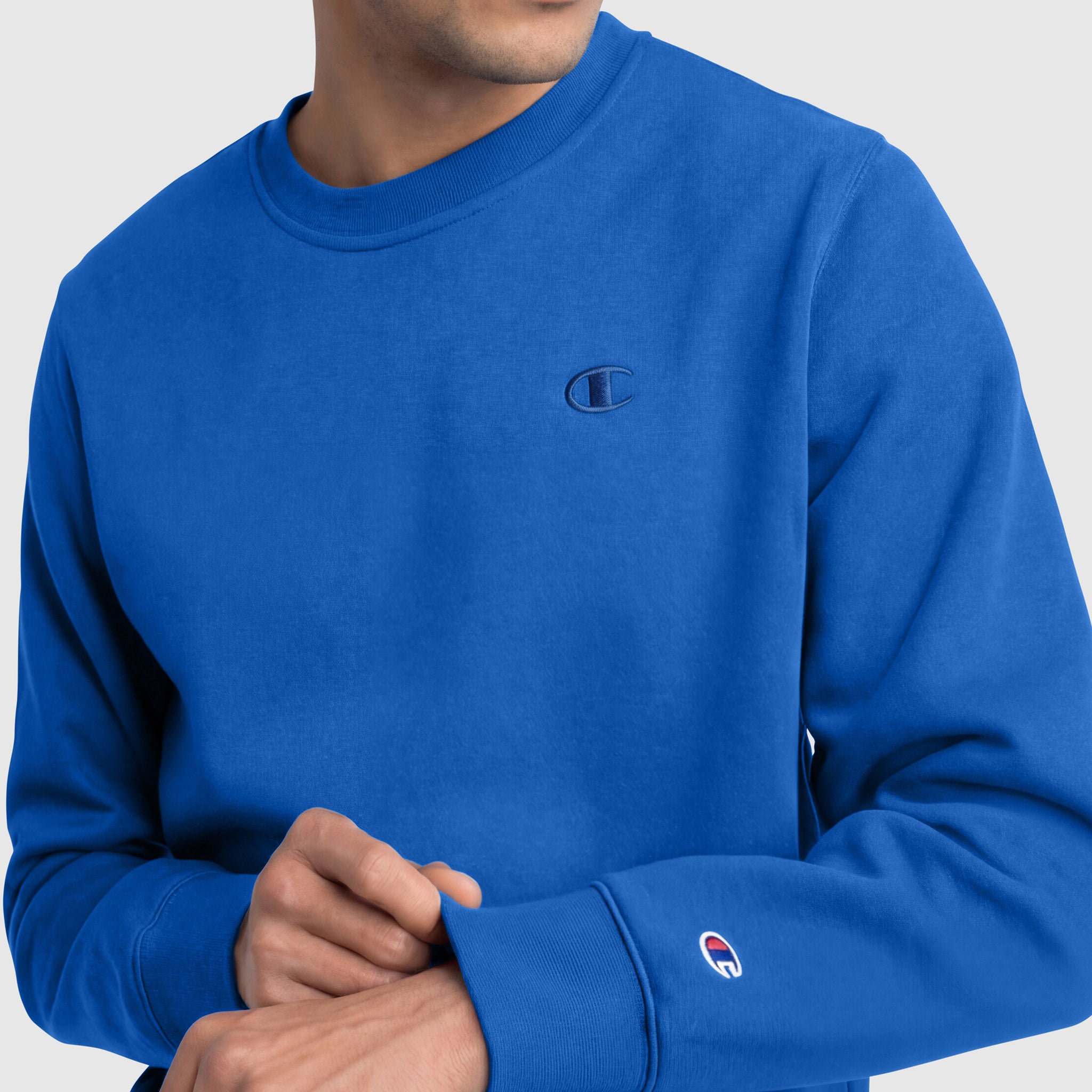 A man adjusts the sleeve cuff of a bright blue Champion Powerblend Crewneck Sweatshirt, C Logo, featuring logo patches on the chest and sleeve, set against a light gray background.