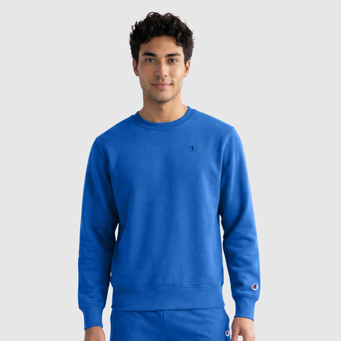 A person with short, curly dark hair is smiling slightly while wearing a bright blue Champion Powerblend Crewneck Sweatshirt with a C Logo, standing against a plain light gray background.