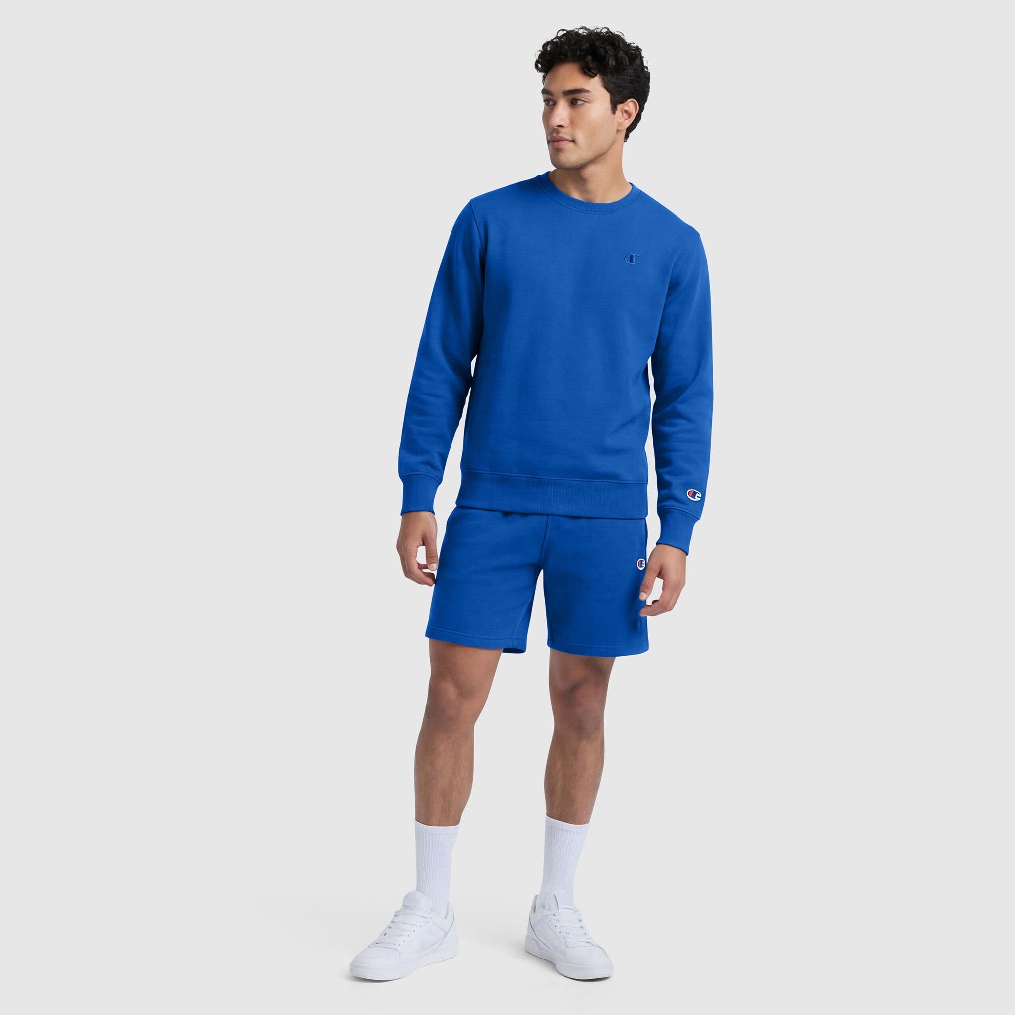 A man wears a Champion Powerblend Crewneck Sweatshirt, C Logo, and matching shorts in royal blue, paired with white socks and sneakers, standing against a plain light gray background.