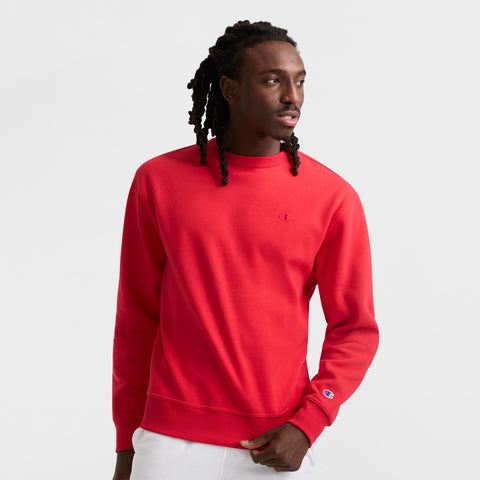 A man with long braided hair is wearing a bright red Champion Powerblend Crewneck Sweatshirt with the C Logo and white pants, standing against a plain white background while looking to his left.