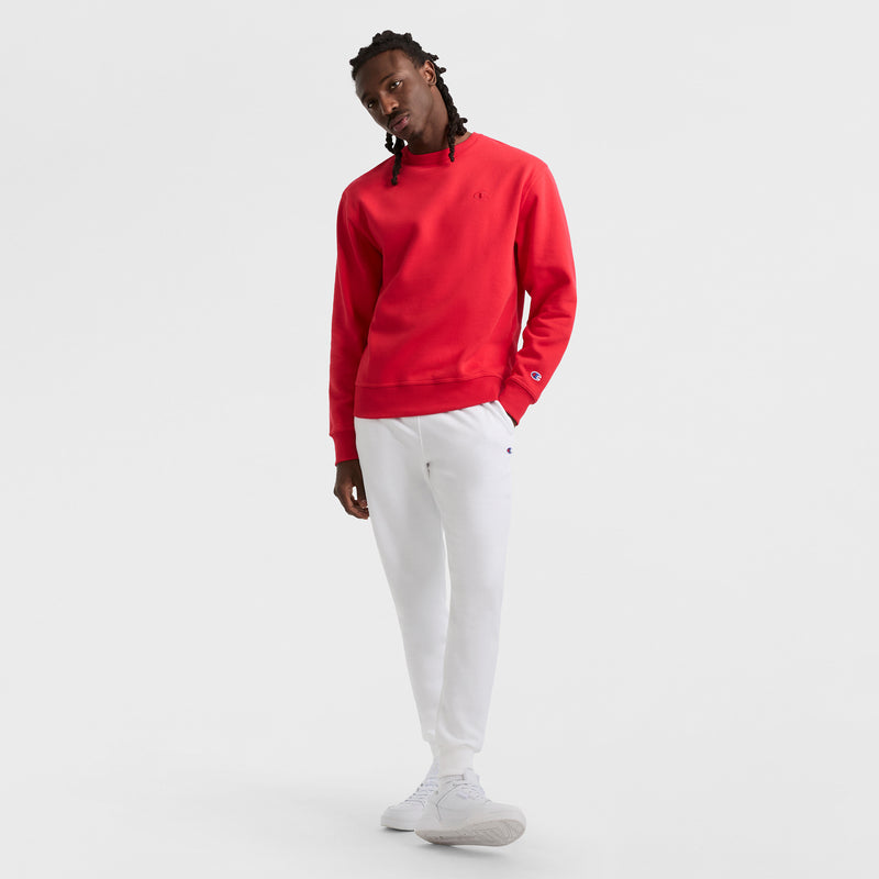 A person stands against a white background wearing a bright red Champion Powerblend Crewneck Sweatshirt (C Logo), white jogger pants, and white sneakers, their head slightly tilted and one hand in their pocket.
