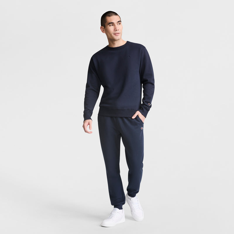 A man stands against a white background wearing the Champion Powerblend Crewneck Sweatshirt with matching sweatpants and white sneakers, looking slightly to the side in a relaxed pose.