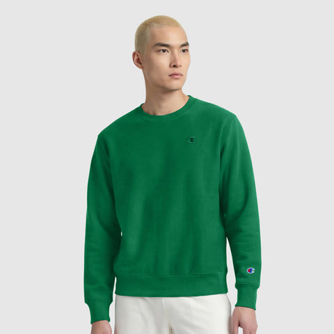 A person with short blond hair wears a bright green Champion Powerblend Crewneck Sweatshirt, C Logo, with white pants, standing against a plain light gray background.