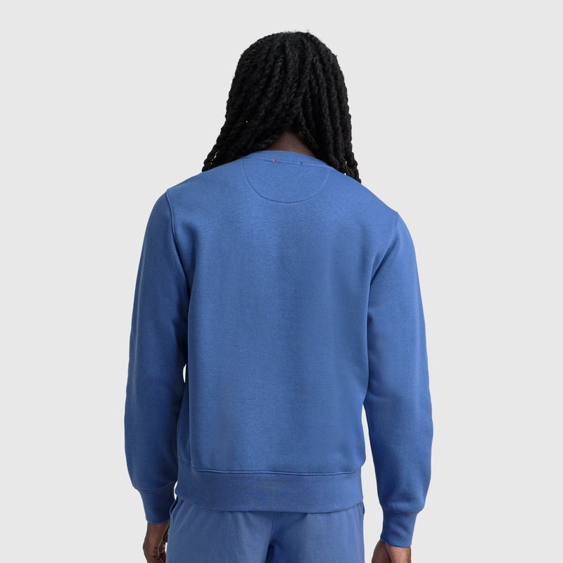 A person with long, black, braided hair is seen from behind wearing a blue Champion Powerblend Crewneck Sweatshirt, C Logo, and matching blue pants against a plain light gray background.