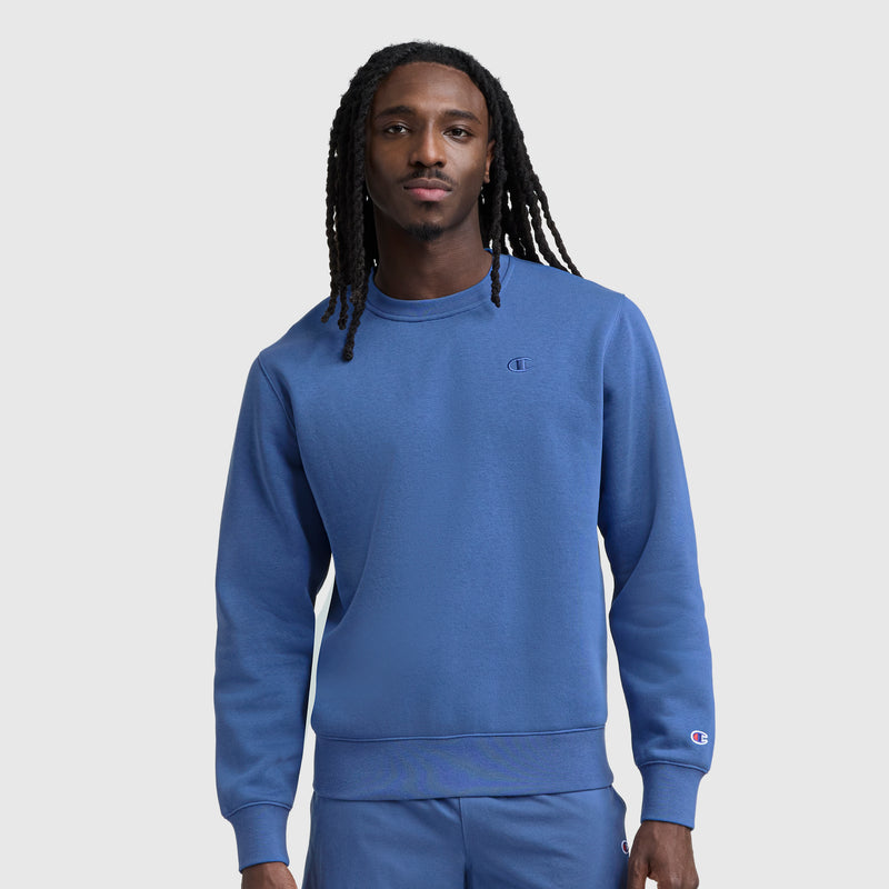 A man with long braided hair wears a blue Champion Powerblend Crewneck Sweatshirt, C Logo, and matching pants, standing against a plain light gray background.