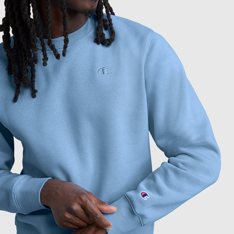 A person with long hair is wearing the Champion Powerblend Crewneck Sweatshirt, C Logo in light blue, featuring the classic athletic fleece design and embroidered Champion logos on the chest and sleeve, set against a plain light background.