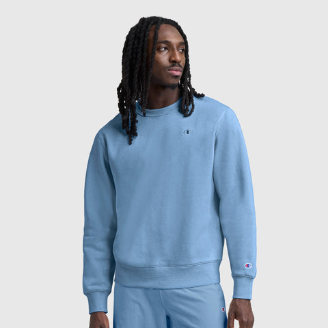 A man with long dreadlocks wears a light blue Champion Powerblend Crewneck Sweatshirt, C Logo, paired with matching pants as he stands against a plain gray background. He faces slightly to his left with a neutral expression.