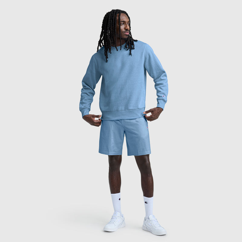A person wearing a light blue Champion Powerblend Crewneck Sweatshirt with the C Logo and blue shorts stands facing slightly to the side. They have long, dark hair and wear white socks and white sneakers against a plain light gray background.