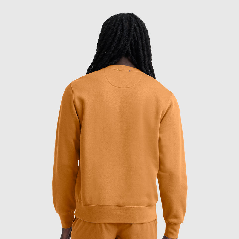 A person with long, dark, braided hair is seen from behind, wearing a Champion Powerblend Crewneck Sweatshirt with C Logo in orange and matching pants against a plain light gray background.