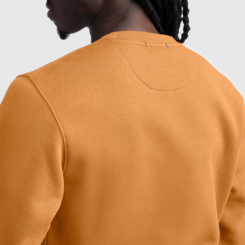 A close-up of a person wearing a mustard yellow Champion Powerblend Crewneck Sweatshirt, C Logo, shown from the back. The fabric texture and neckline details are visible, and the person has dark braided hair.