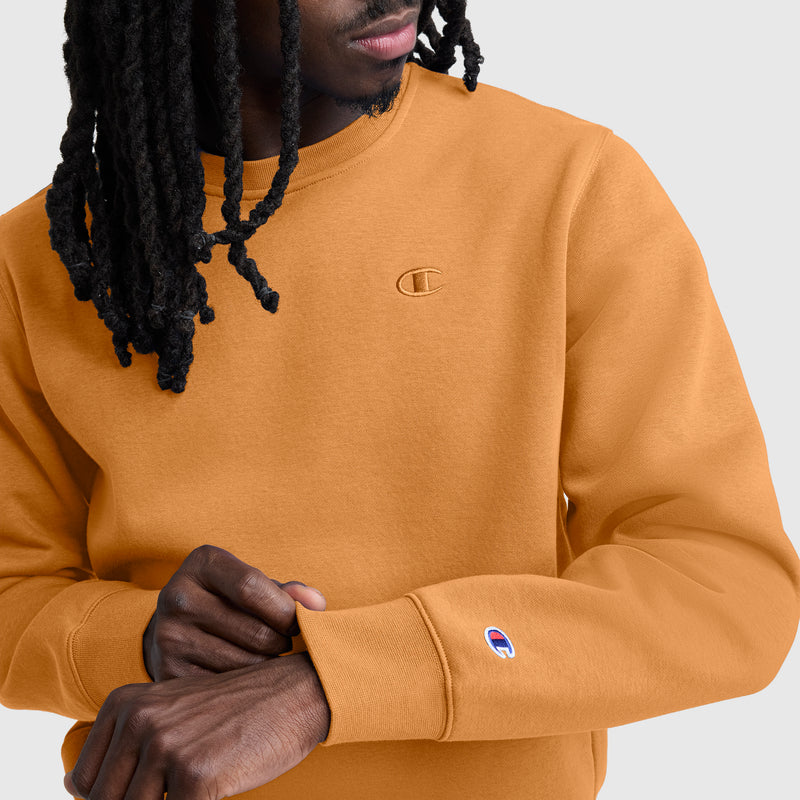 A person wearing a mustard yellow Champion Powerblend Crewneck Sweatshirt, C Logo, adjusts their sleeve. This men’s fleece top showcases the iconic Champion logo on the chest and sleeve. The background is a plain light gray.