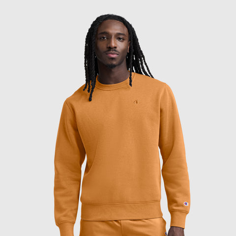 A man with long braids stands against a plain light gray background wearing a Champion Powerblend Crewneck Sweatshirt, C Logo, in orange, paired with matching pants.