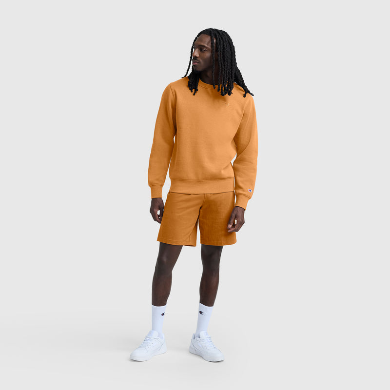 A man with long hair is wearing a matching orange Champion Powerblend Crewneck Sweatshirt with C Logo and shorts set, paired with white crew socks and white sneakers, standing against a plain light gray background.