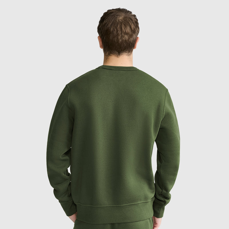 A person with short brown hair is shown from the back, wearing a plain dark green Champion Powerblend Crewneck Sweatshirt, C Logo, along with matching pants, standing against a light gray background.