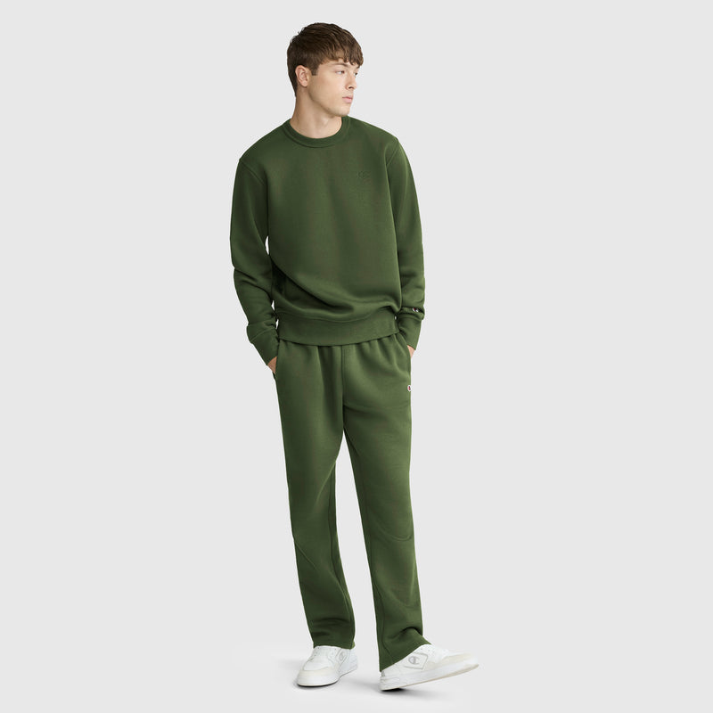 A young man is wearing a Champion Powerblend Crewneck Sweatshirt, C Logo, in olive green along with coordinating sweatpants. He stands with his hands in his pockets, looking to the side, and completes the look with white sneakers against a plain light gray background.