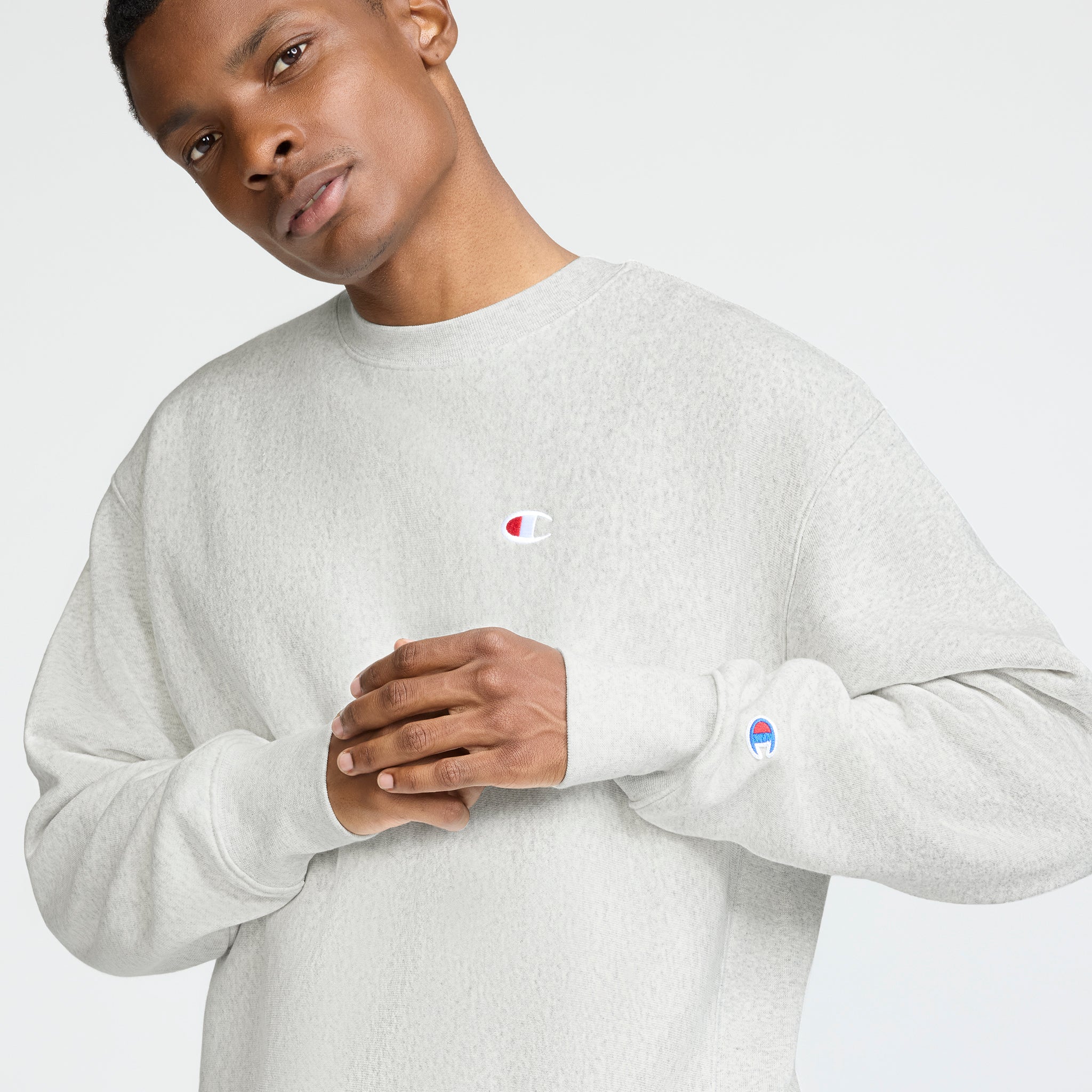 A person wears a light gray Champion Reverse Weave Crewneck Sweatshirt, C Logo, standing with hands clasped and looking upward. The heavyweight fleece features Champion’s C logo on the chest and sleeve, set against a light background.