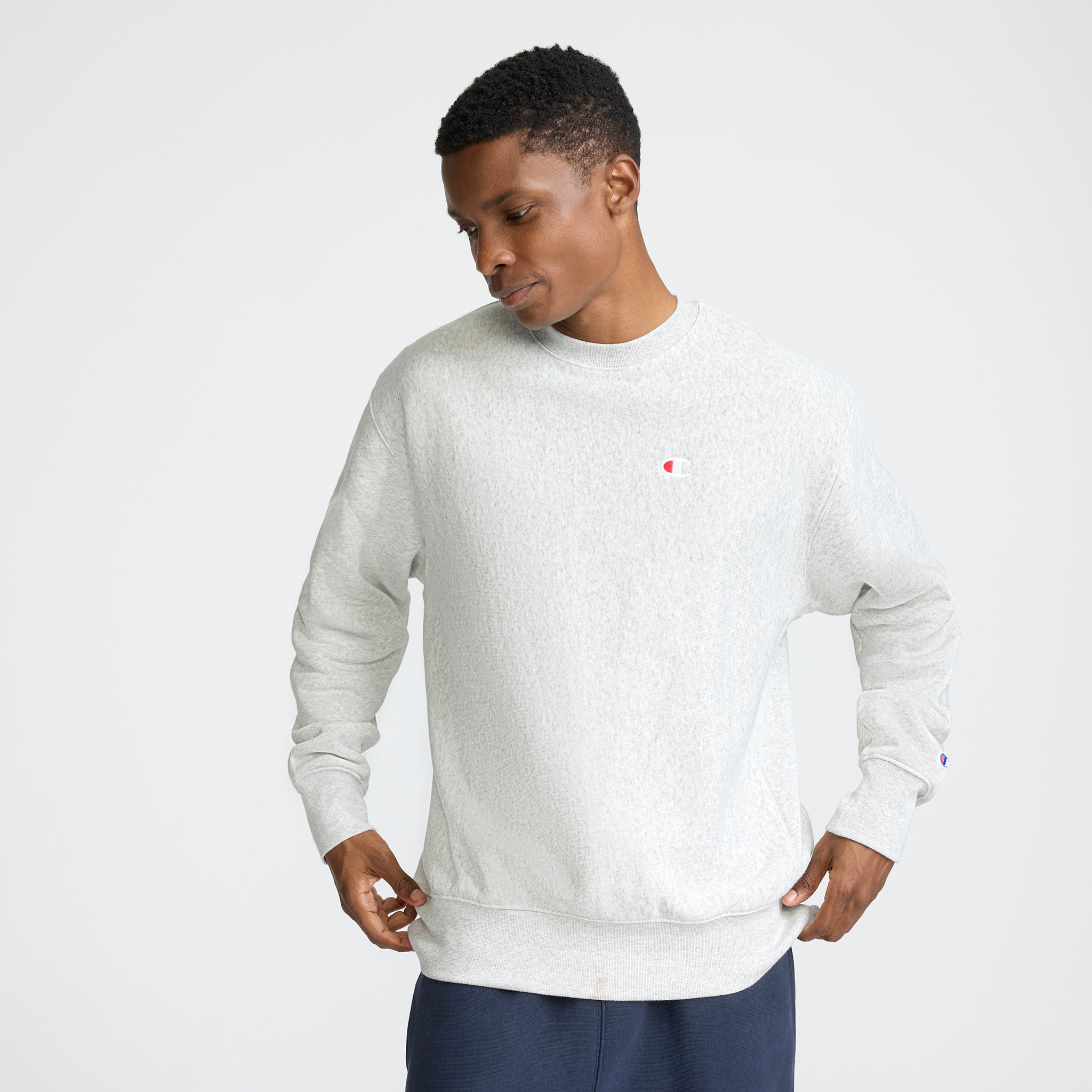 A man stands against a plain background, looking down while holding the sides of his light gray Champion Reverse Weave Crewneck Sweatshirt, featuring the C logo on the chest. He pairs it with dark pants.