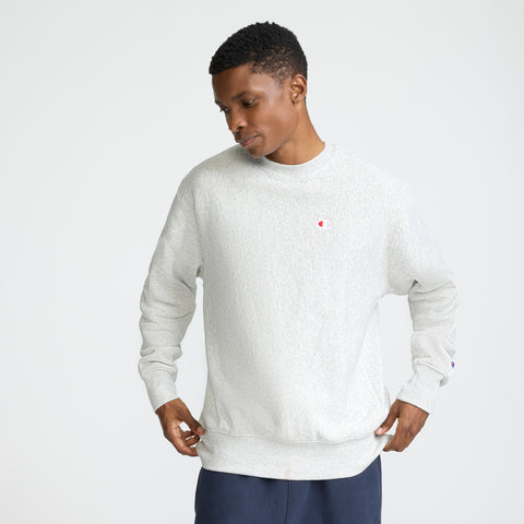 A man stands against a plain background, looking down while holding the sides of his light gray Champion Reverse Weave Crewneck Sweatshirt, featuring the C logo on the chest. He pairs it with dark pants.