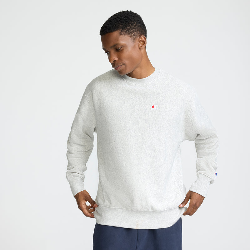 A man stands against a plain background, looking down while holding the sides of his light gray Champion Reverse Weave Crewneck Sweatshirt, featuring the C logo on the chest. He pairs it with dark pants.