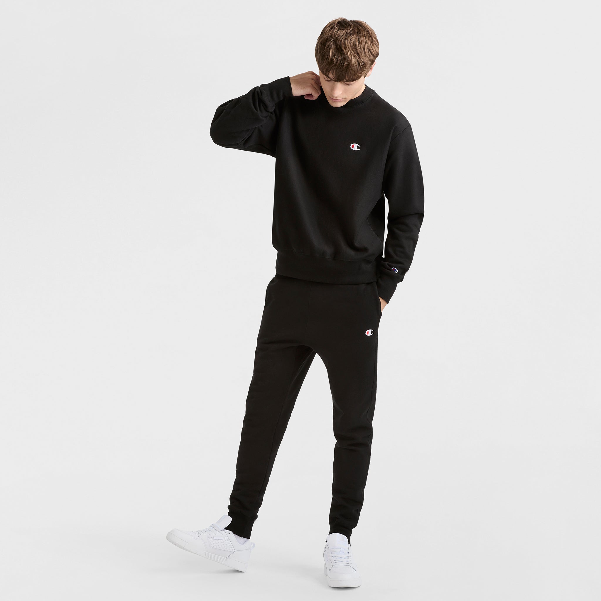 A person wearing a Champion Reverse Weave Crewneck Sweatshirt, C Logo, and matching black sweatpants with white sneakers stands against a plain white background, looking down with one hand touching their neck.