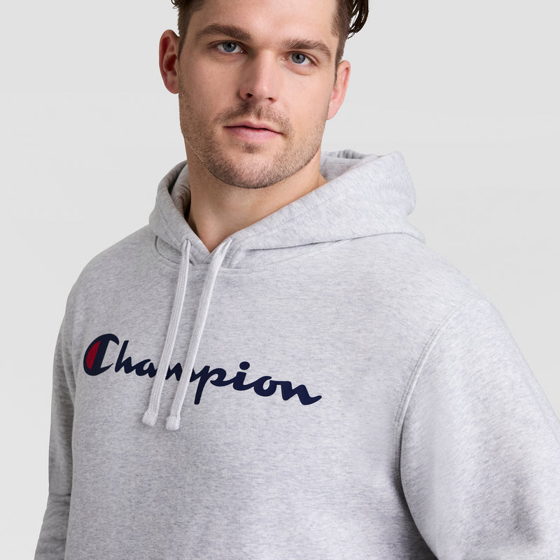A man wearing a light gray Champion Powerblend Hoodie, Full Script Logo - Big & Tall, featuring "Champion" in navy blue and red on the front, stands against a plain light background.
