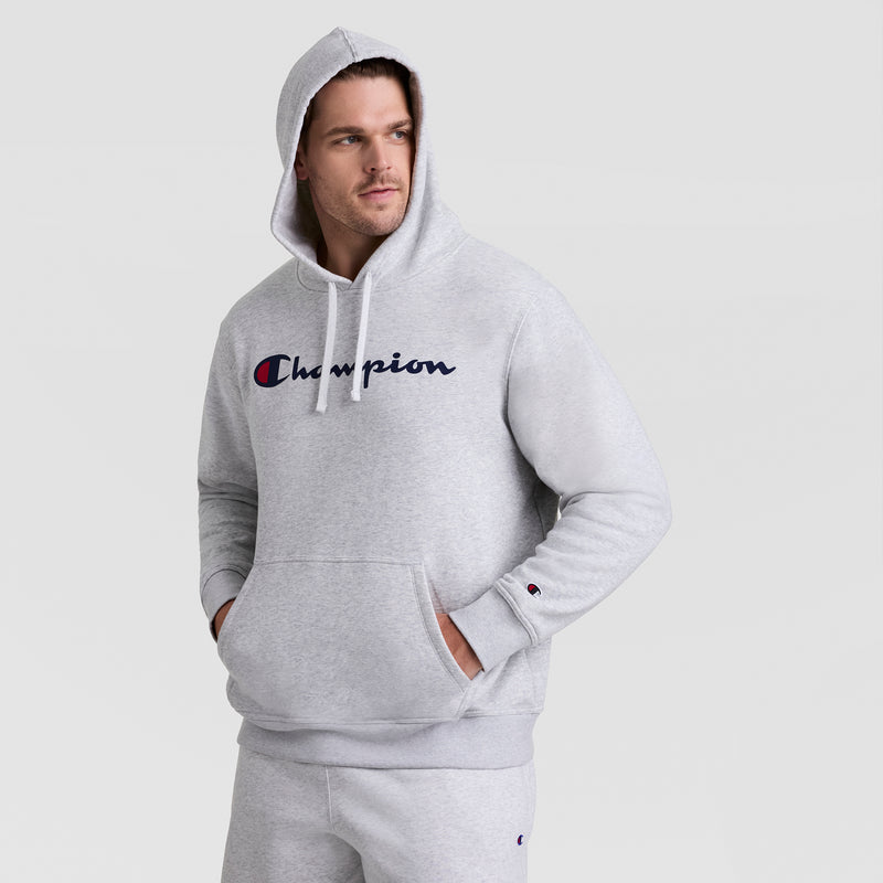 A man wearing a light gray Champion Powerblend Hoodie, Full Script Logo - Big & Tall, with a front pocket and the Champion logo across the chest stands with one hand in his pocket, looking to the side.