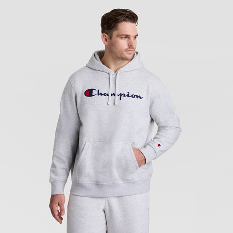 A man wears the Champion Powerblend Hoodie, Full Script Logo - Big & Tall in light gray with matching sweatpants, standing against a white background and looking slightly right with one hand in his pocket.