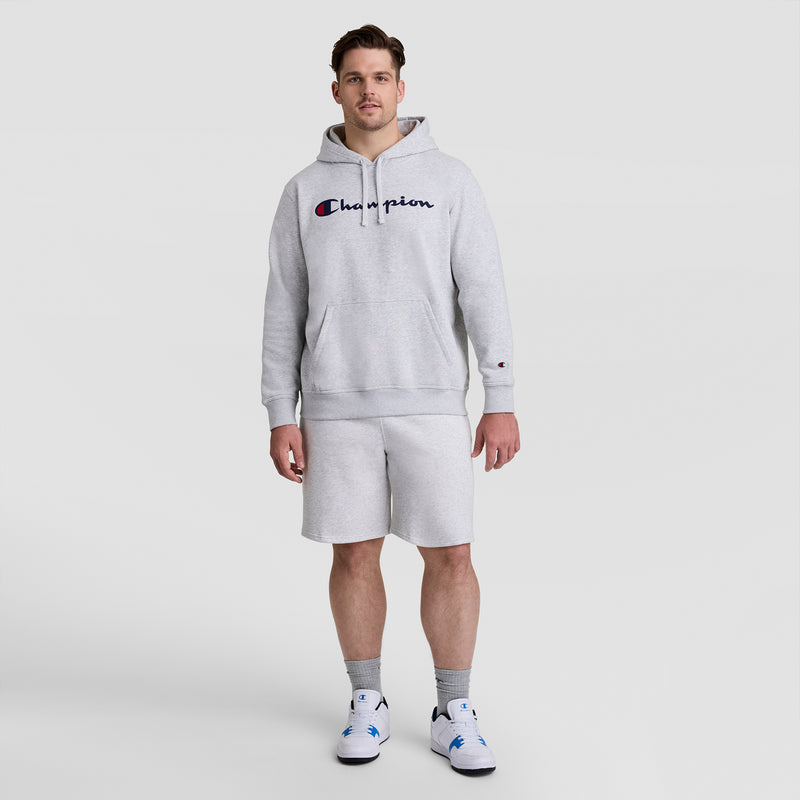 A man stands against a plain light background wearing a Champion Powerblend Hoodie, Full Script Logo - Big & Tall in light gray, matching shorts, gray socks, and white sneakers with blue accents.