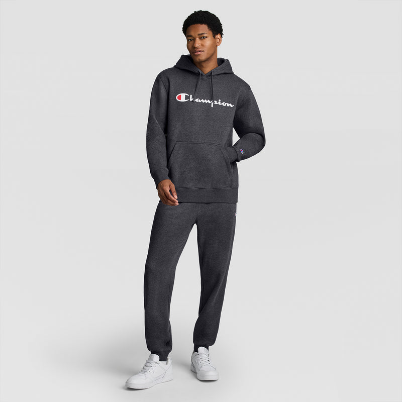 A person wearing a dark gray Champion Powerblend Hoodie, Full Script Logo, with matching sweatpants stands against a plain light gray background, white sneakers completing the outfit.