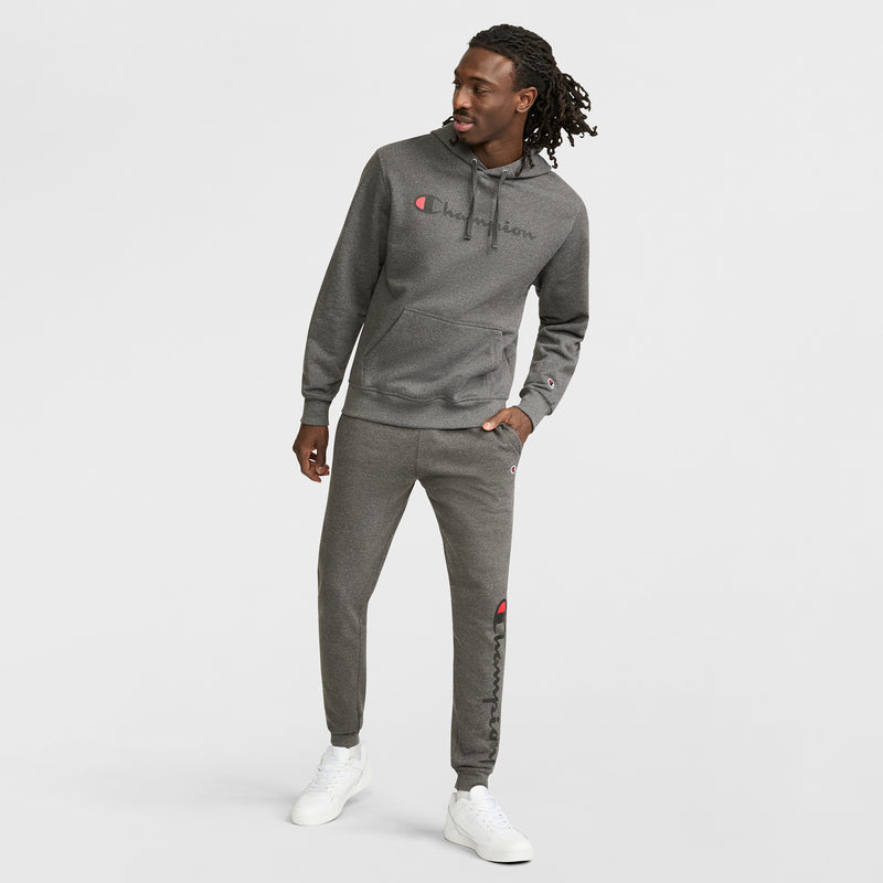 A man with long hair wears a Champion Powerblend Hoodie, Full Script Logo, in grey with matching sweatpants and white sneakers, standing in a relaxed pose against a plain white background.
